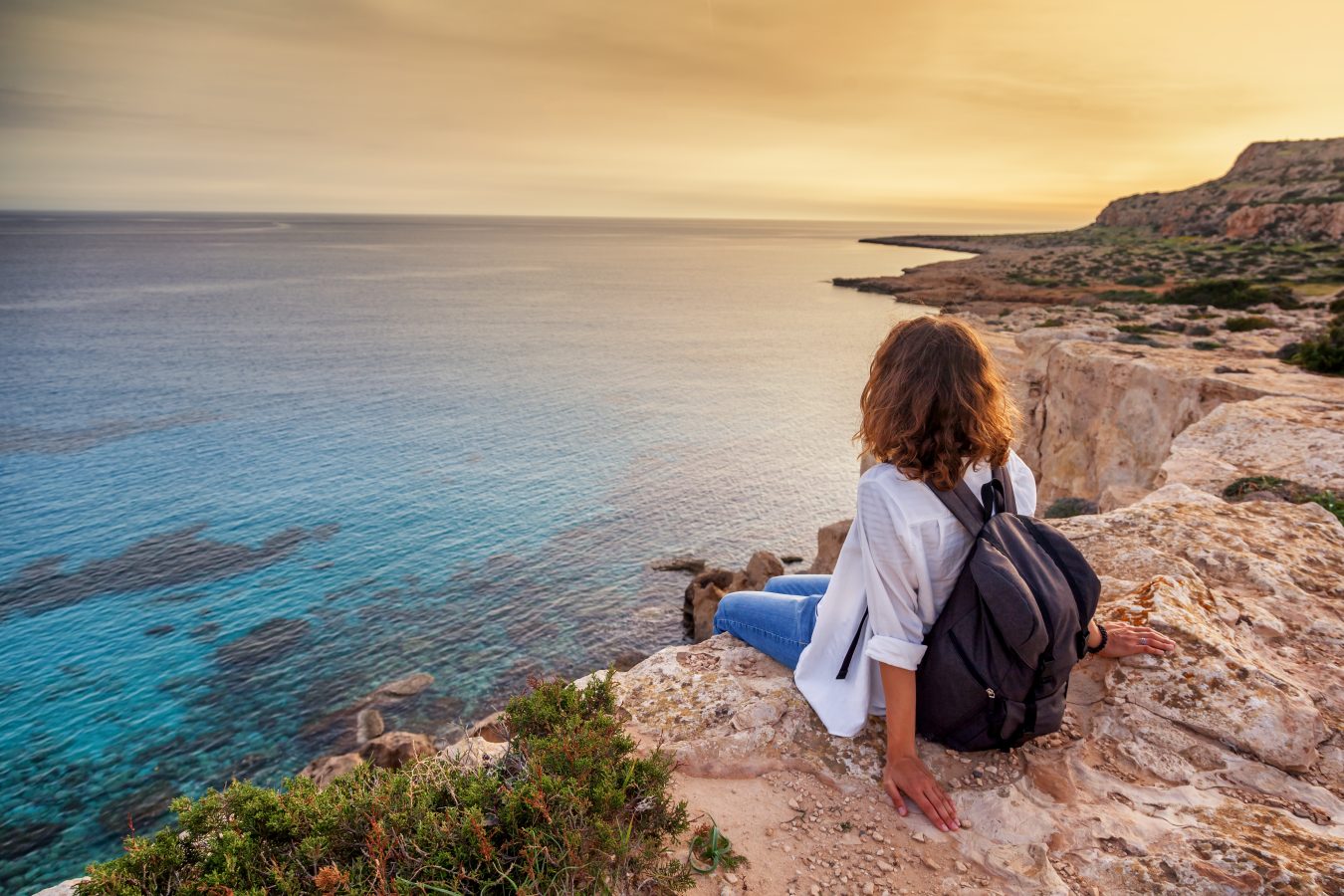 A young woman traveler watches a sunset on the rocks of a cliff overlooking the sea in Cyprus - a popular and cheap Europe destination for summer travel.