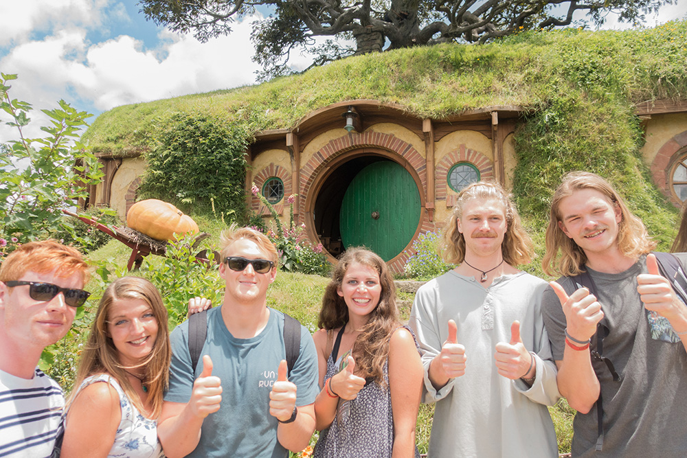 Group of young adults posing with thumbs up in front of a hobbit looking home on an adventure travel trip.