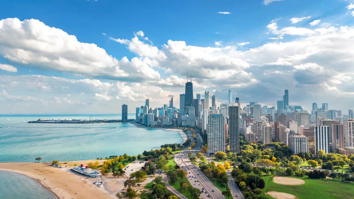 Chicago's skyline to the right of the image with a light blue summertime Lake Michigan to the left on a sunny day. Chicago is one of the cheapest places to fly right now.