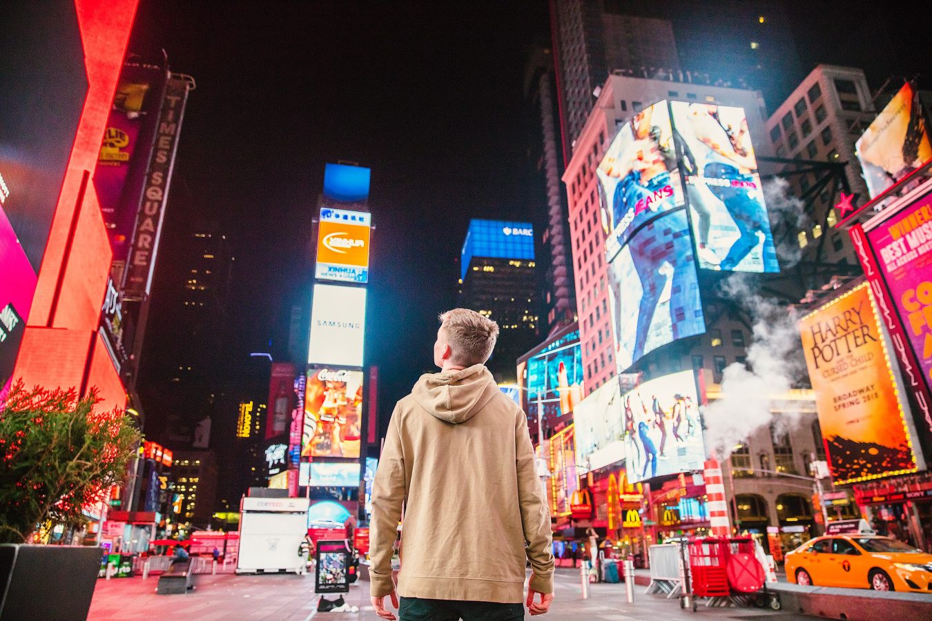 A young guy stands in a hoodie in Times Square in New York City at night.