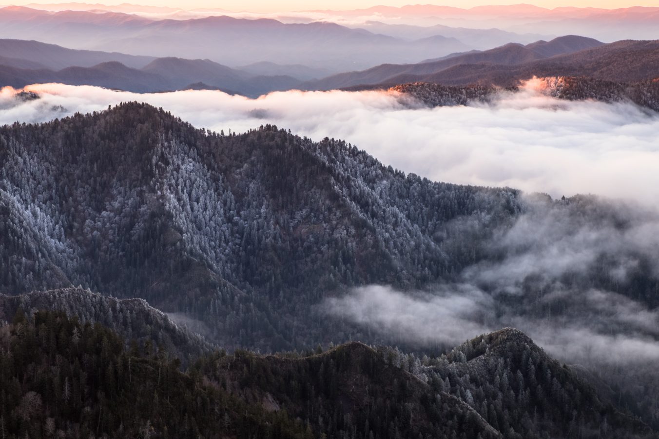 From a viewpoint high in the Great Smokey Mountains National Park in winter stretches a line of mountain ridges to the horizon. It is sunrise and the sky is orange. Low clouds fill the valleys. Misty and foggy