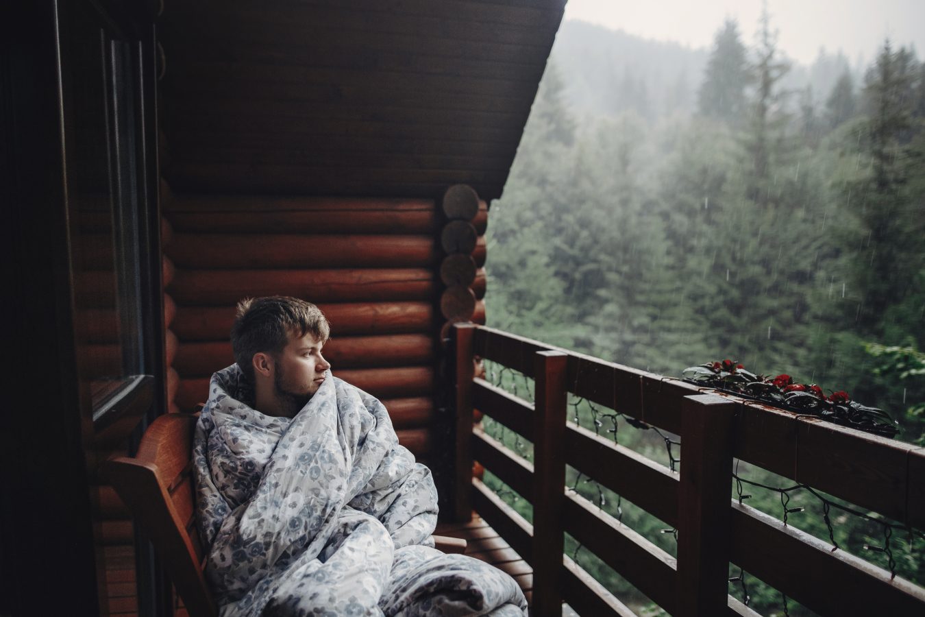 Man in blanket sitting on cabin porch in the rainy woods.