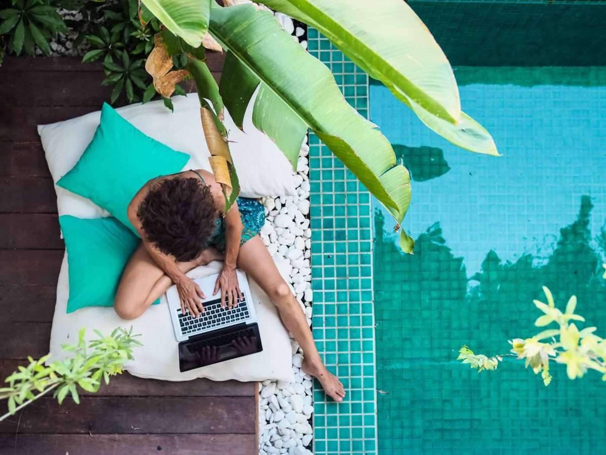 A digital nomad is sitting on a lounger, working on a laptop by the pool