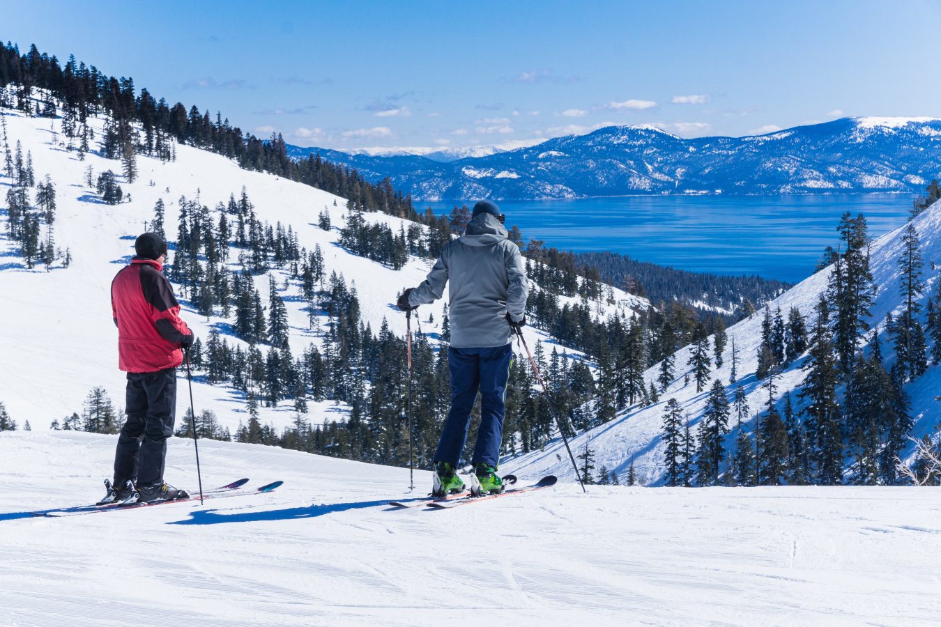 Two guys on skis look out over Lake Tahoe and the surrounding mountains on a sunny spring day.