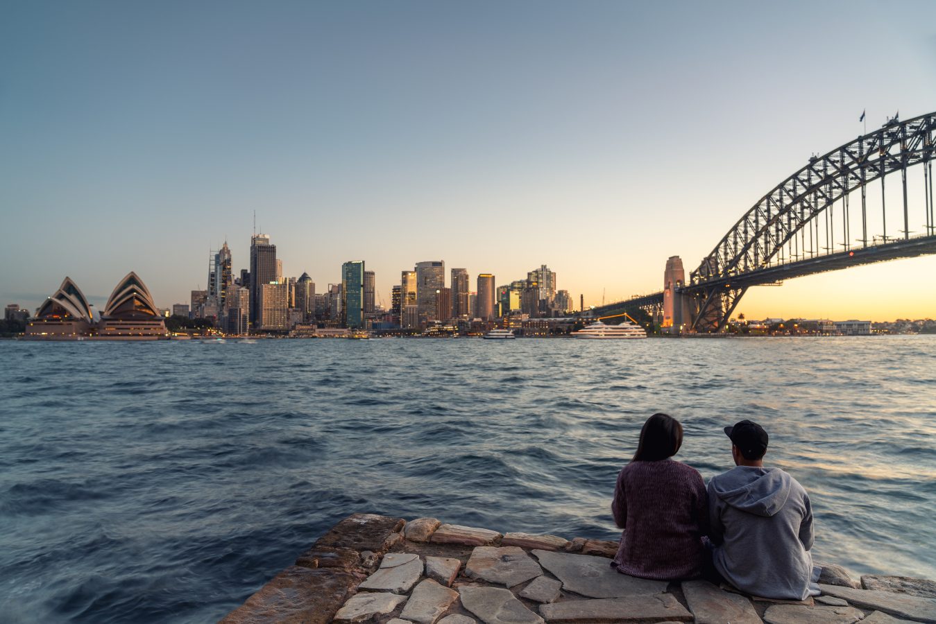Girl and guy sit at sunset on the water overlooking Sydney skyline and Sydney Opera House in Sydney, New South Wales, Australia.