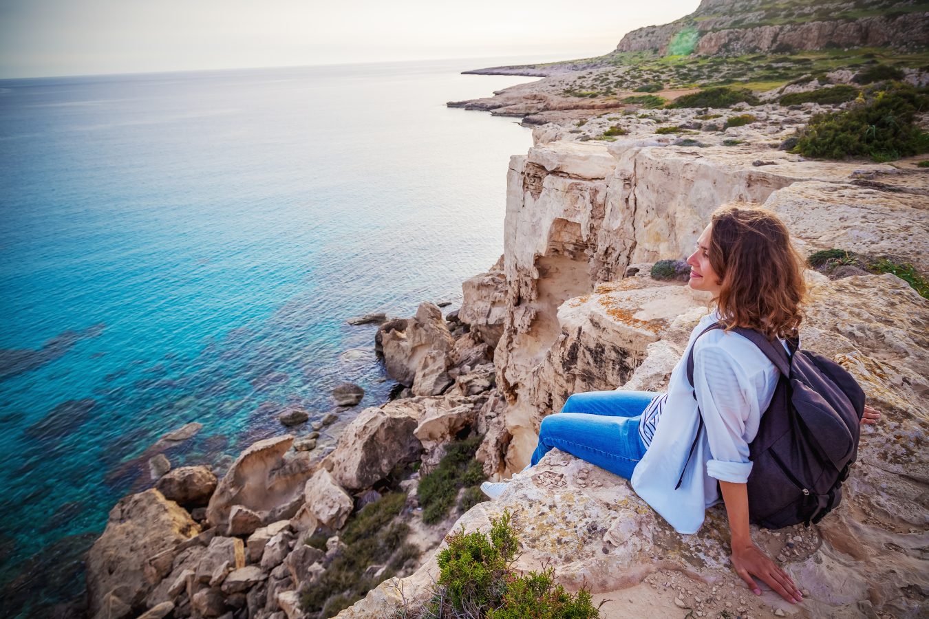 Young woman sits on rocky cliff and watches the ocean in Greece, a popular destination for cheap travel.