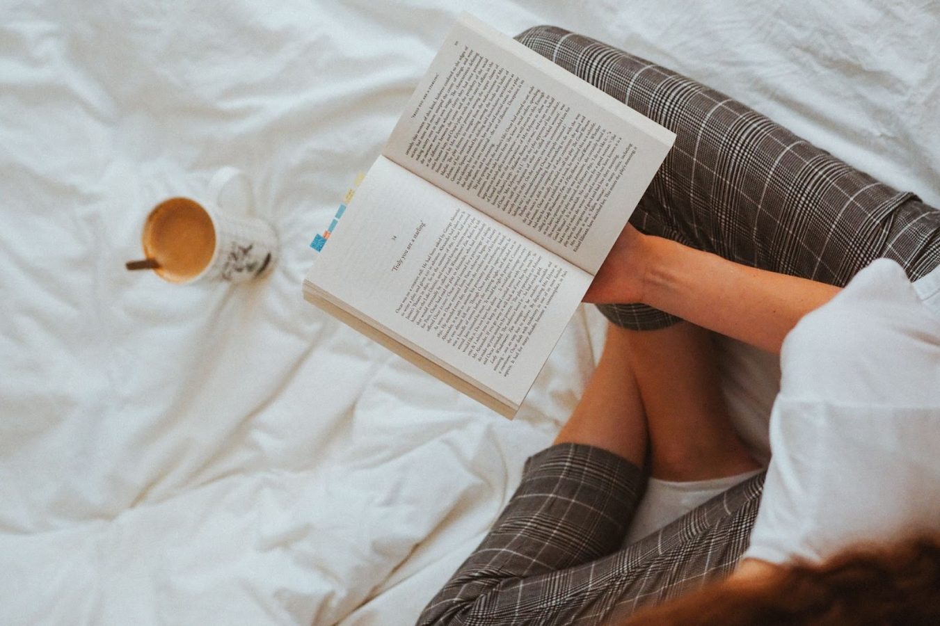 Top-down view of a person reading a book while sitting cross legged on white sheets with a mug of tea.
