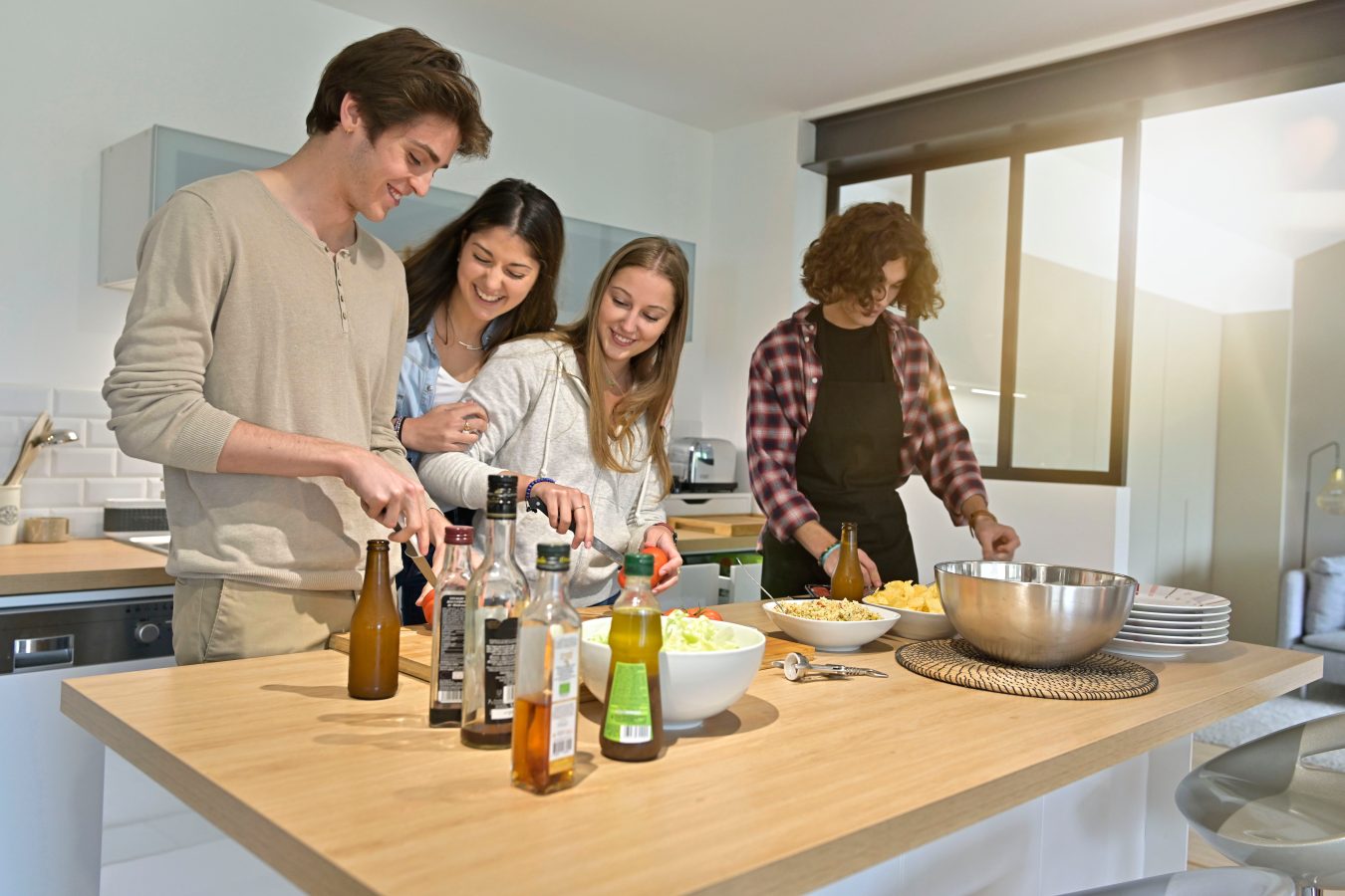 Four roommates cooking dinner together in their apartment kitchen, helping them to save money for a trip by not eating out.