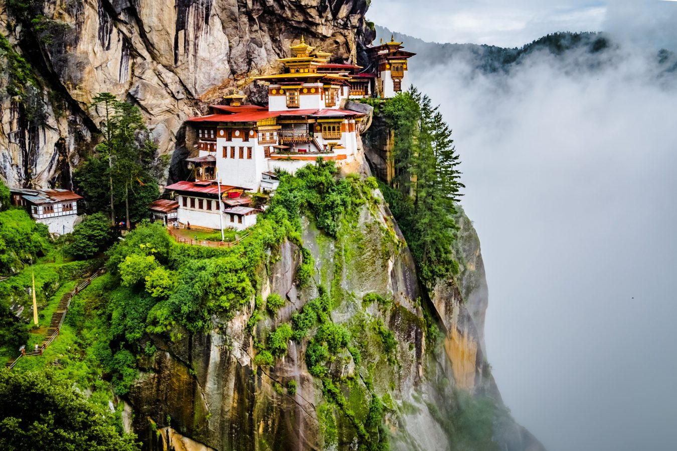View of Tiger's nest monastery in Bhutan which sits on the side of a mountain cliff as clouds of mountains are in the valley beside.