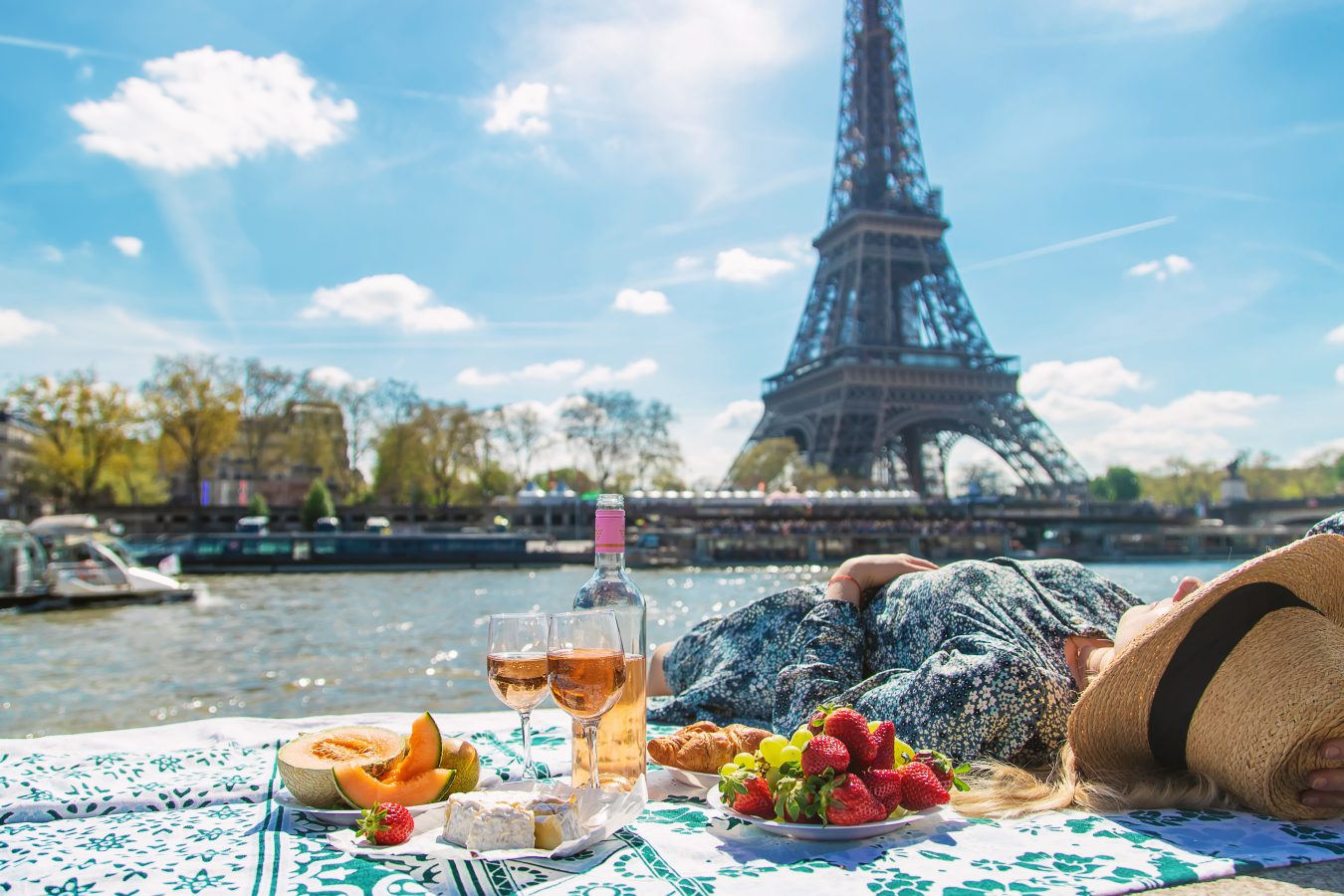 A woman lays on a blanket with a picnic spread next to here along the river in front of the 
Eiffel Tower in Paris.