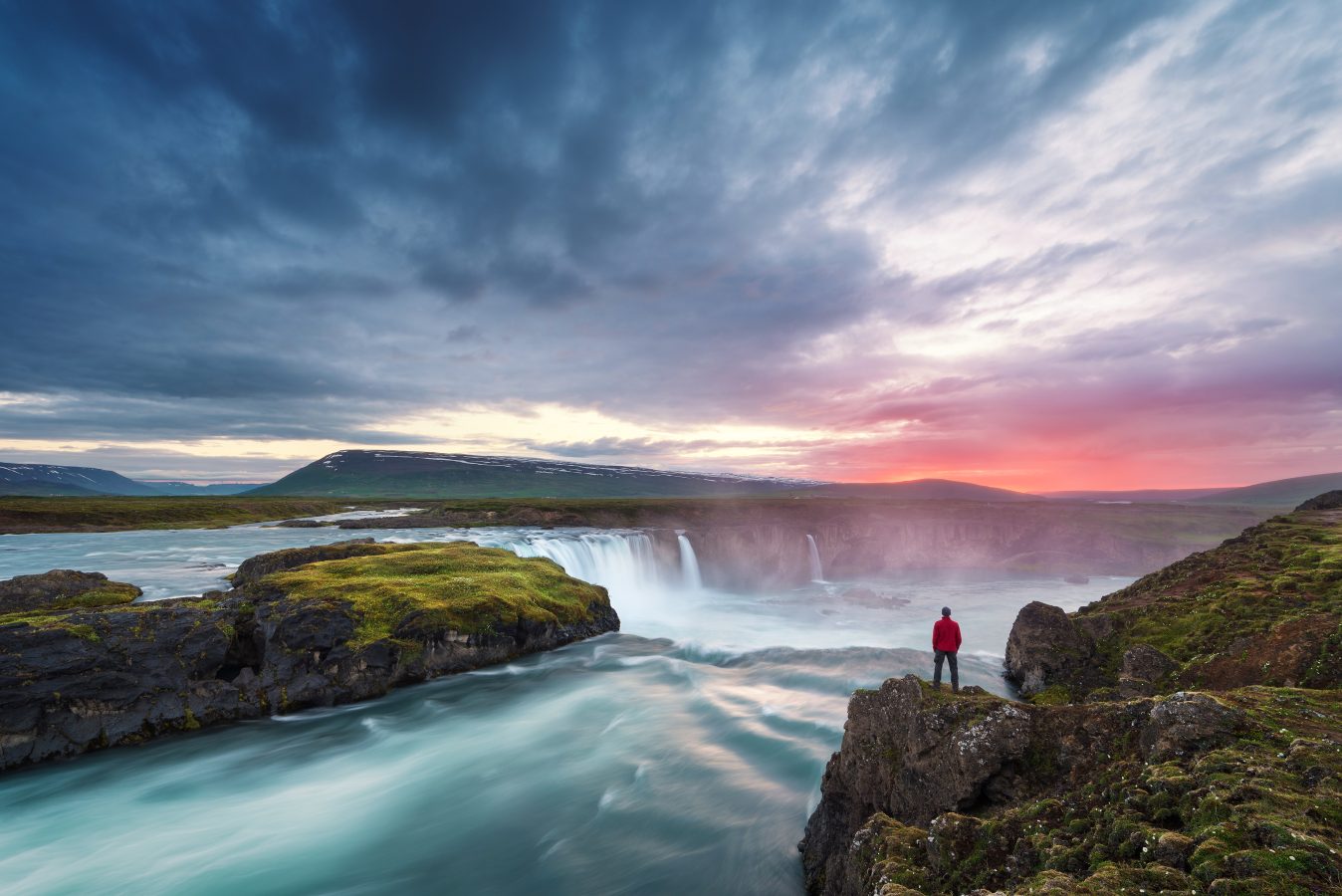 Godafoss waterfall in Iceland with a young guy standing cliffside during sunrise.