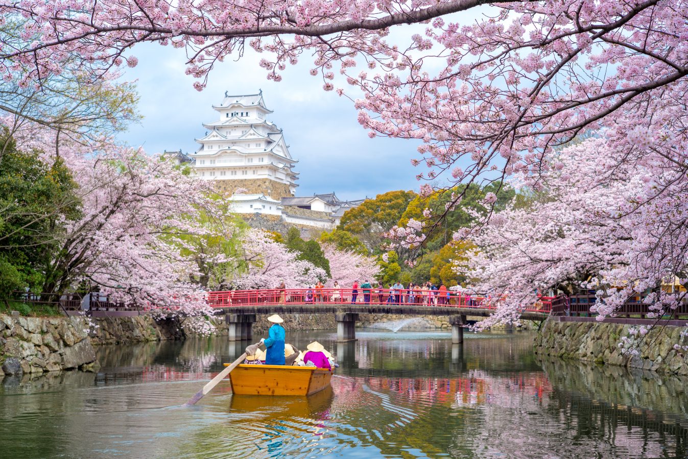 A boat rows along a river in Japan during the spring with cherry blossoms in full bloom and Himeji Castle in the background.