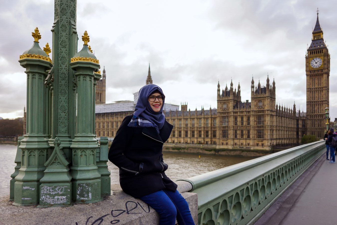 F1 visa student traveler wearing a hijab sits on a beach in London with Big Ben in the background.