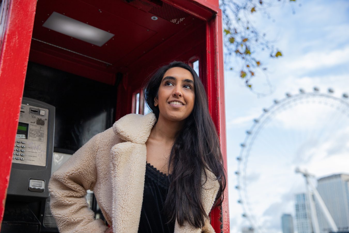F1 visa student traveler poses in London telephone box with London Eye in the background.